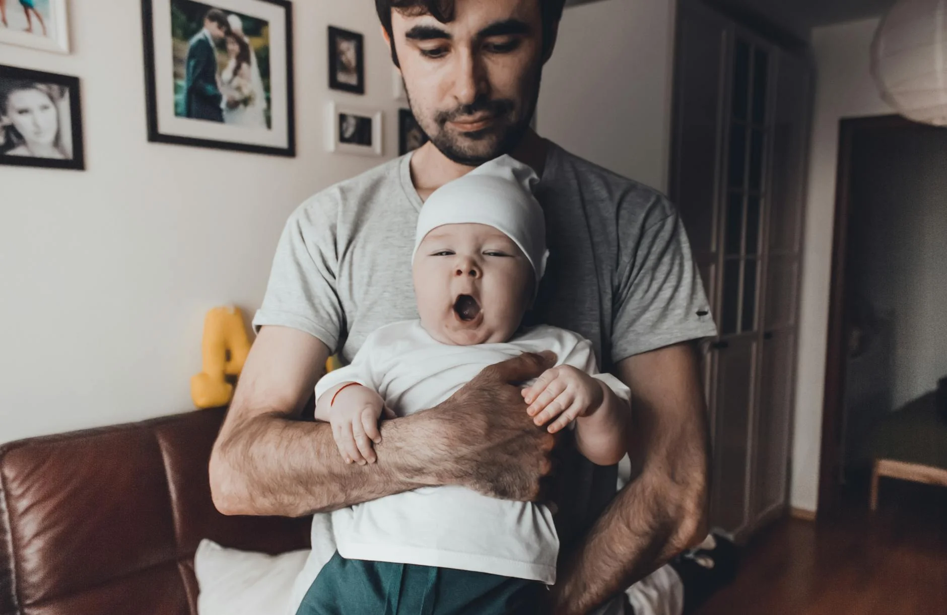 A father lovingly holds his yawning baby inside a warmly lit home, highlighting father-child bonding.