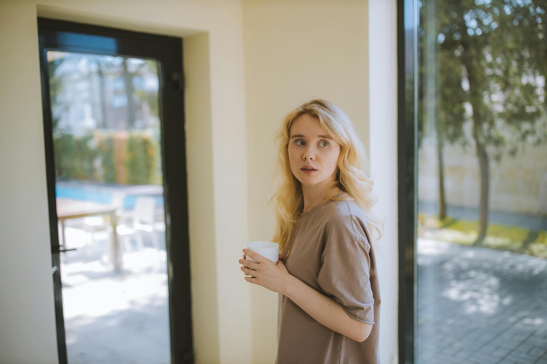 Blonde woman holding a cup near glass doors, looking back indoors.