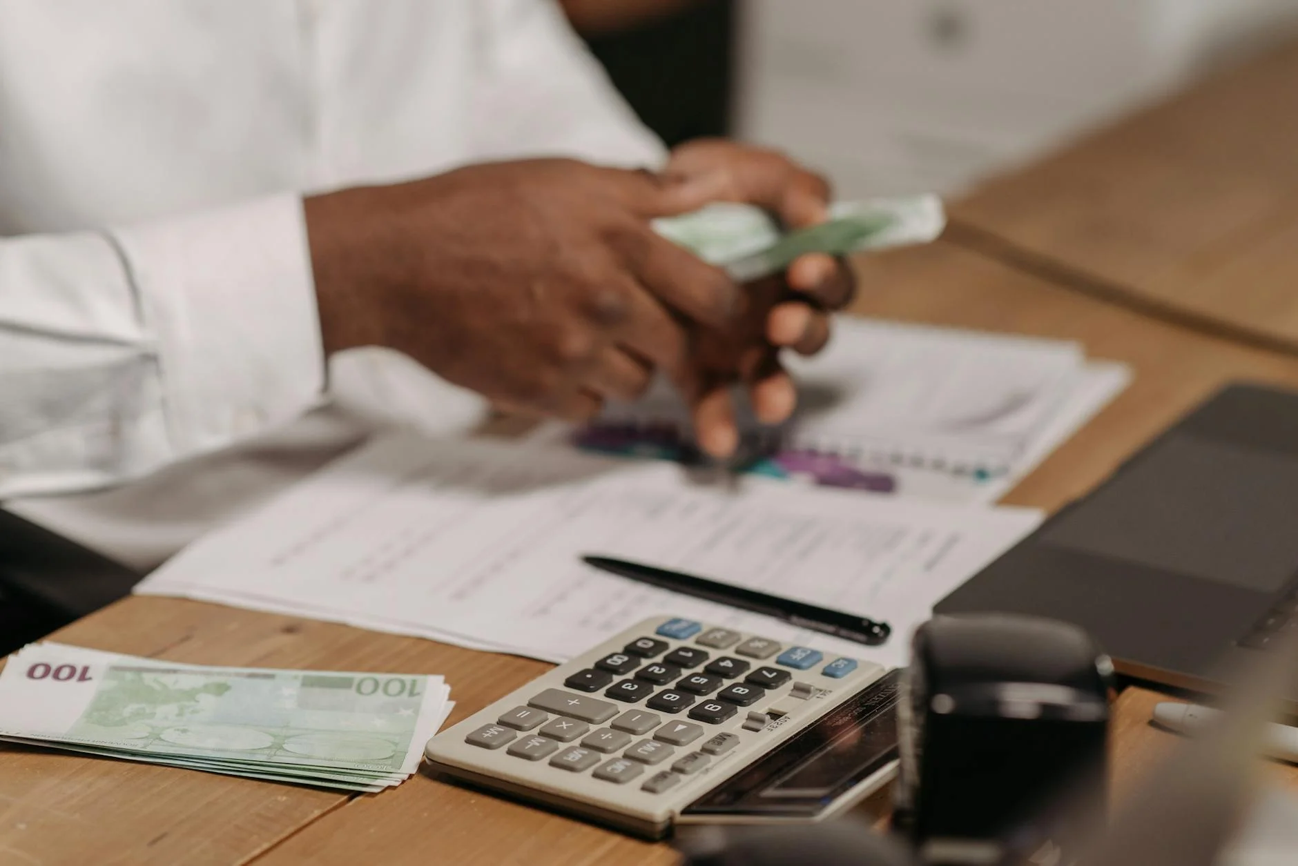 Hands counting euro bills on a wooden desk with calculator, financial documents, and laptop nearby.