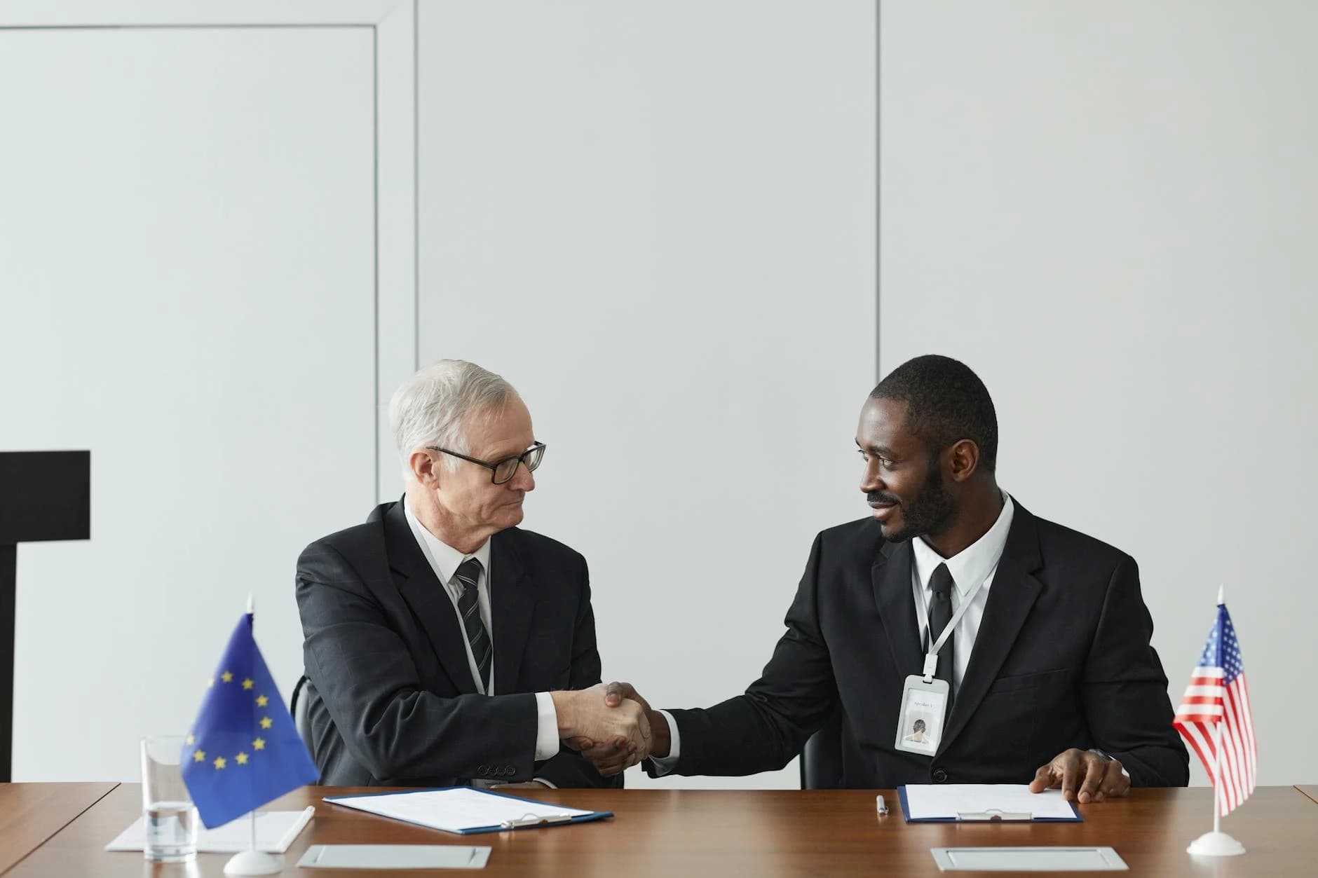 Two men in suits, representing the European Union and the United States, shake hands across a conference table in a professional, neutral office setting. The interaction captures a sense of formal collaboration and diplomatic partnership between the two representatives.