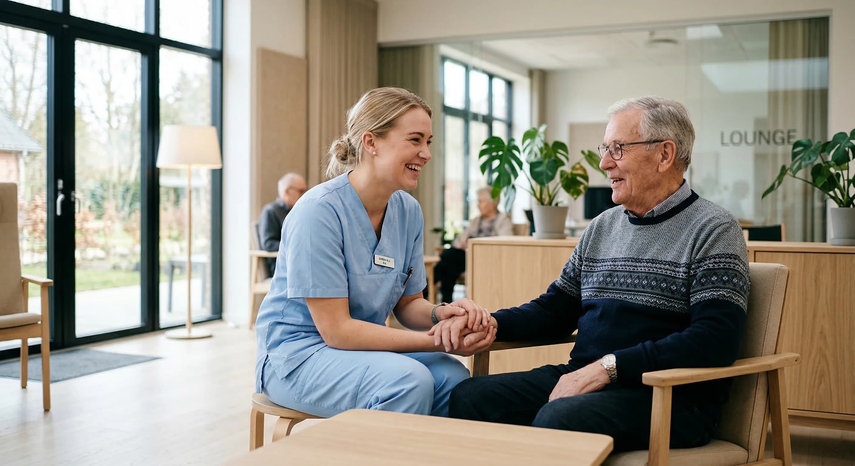 A high-quality, candid photograph of a female healthcare worker in light blue scrubs interacting warmly with an elderly citizen in a bright, modern interior. The room features minimalist Scandinavian