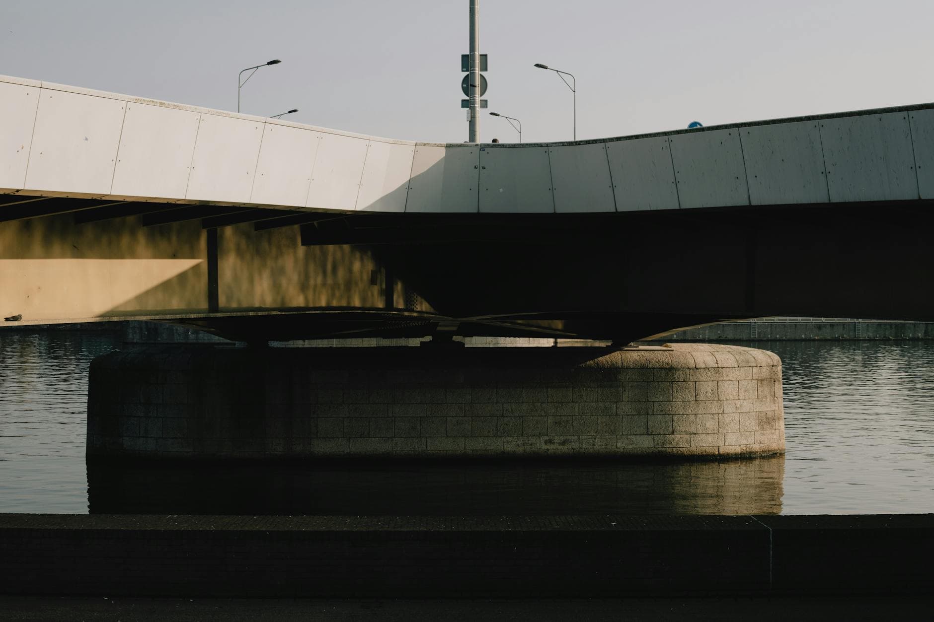 A shadowed section of a modern bridge over a tranquil river in Maastricht at sunset.