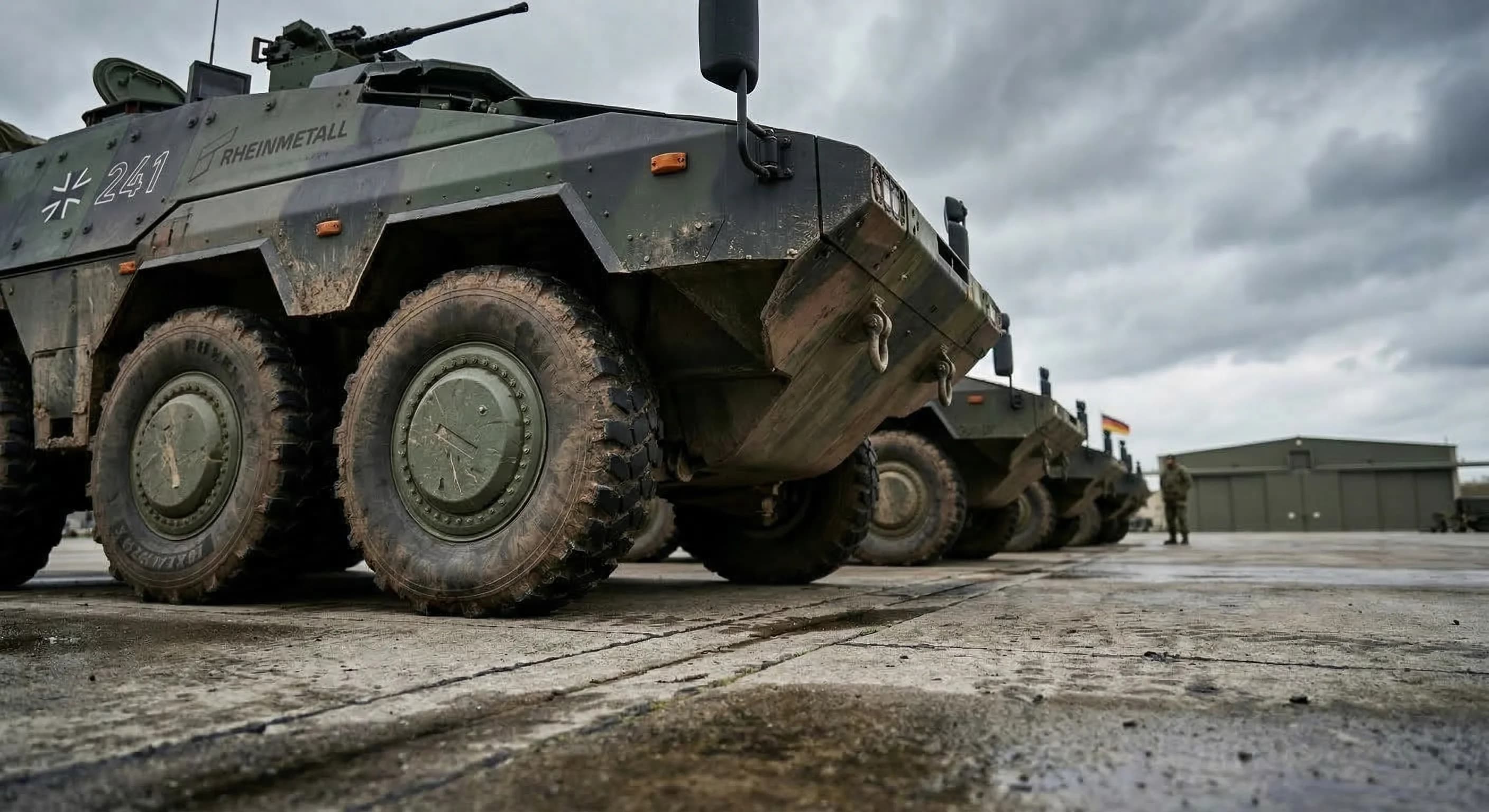 A wide-angle, low-angle shot of a row of modern armored infantry vehicles parked on a concrete airfield under a dramatic, overcast sky. The lighting is soft and natural, highlighting the matte green a