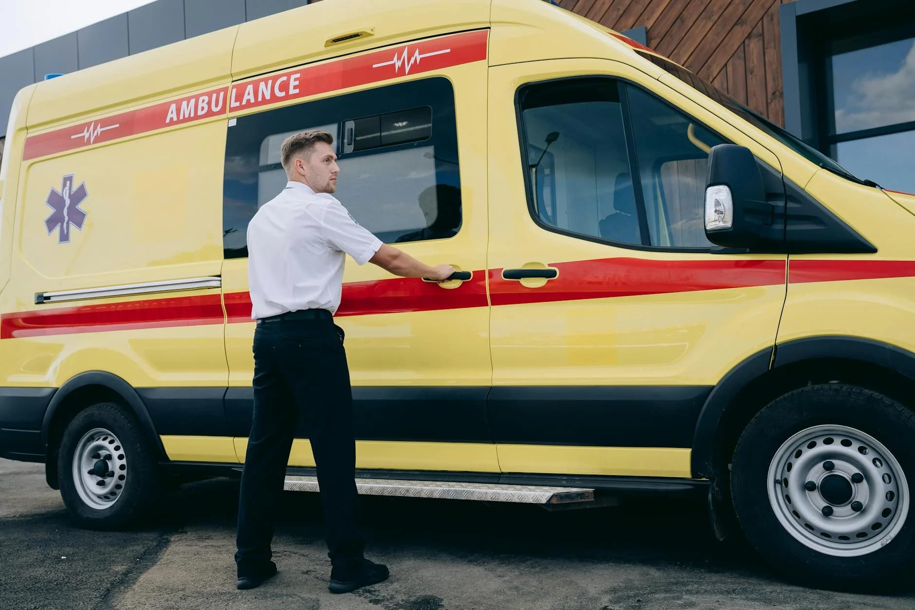 A paramedic in uniform opens the door of a yellow ambulance outside a facility.