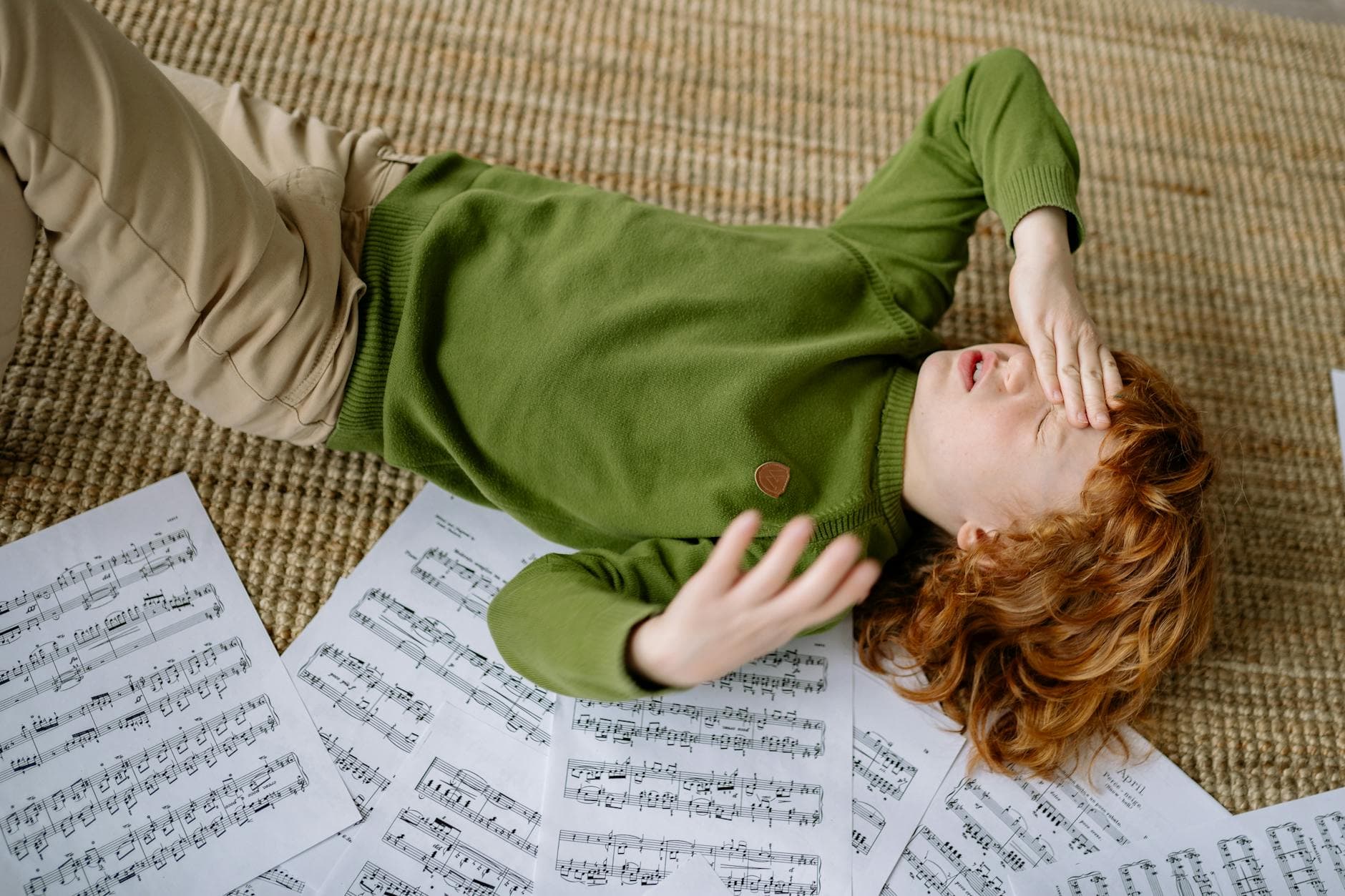A child lying on a rug surrounded by music sheets, expressing frustration.