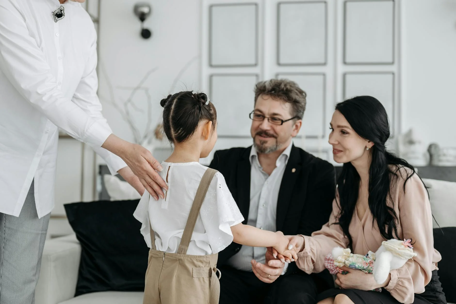 In a bright, modern interior, a young girl is warmly welcomed by a couple, creating an atmosphere of gentle anticipation and emotional connection as she reaches out to hold the woman's hand. An adult, likely an adoption professional, stands beside the child with a reassuring hand on her back, capturing a poignant moment of beginning a new family journey.