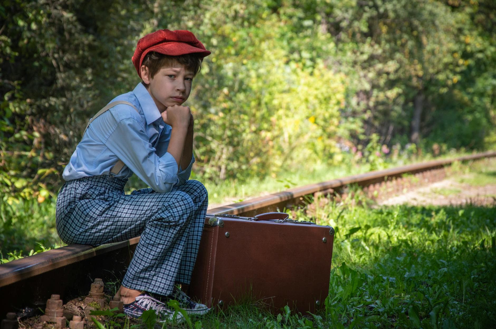 A teenager in vintage attire sits by railway tracks, contemplating travel with a suitcase.