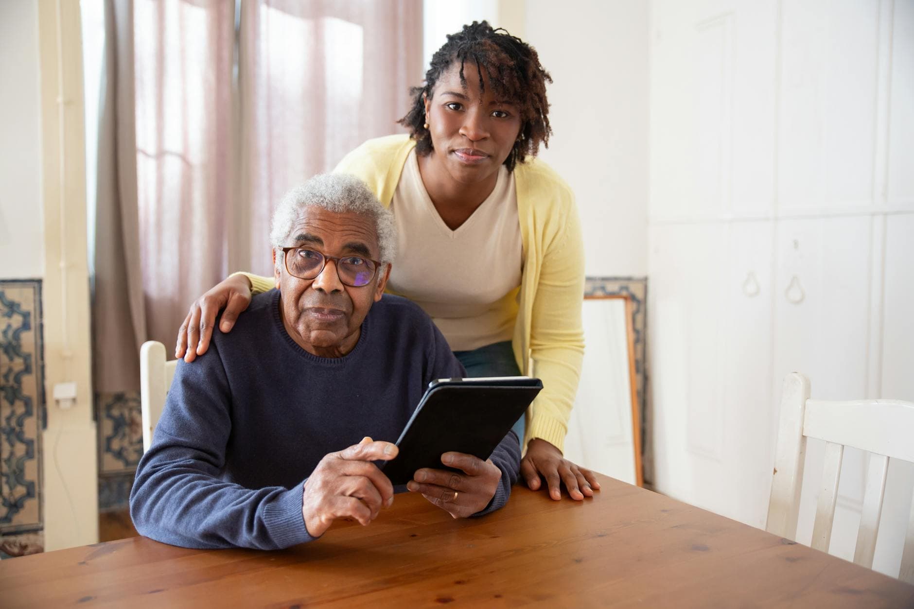 A senior man with eyeglasses and caregiver using a tablet at home, focusing on care and companionship.