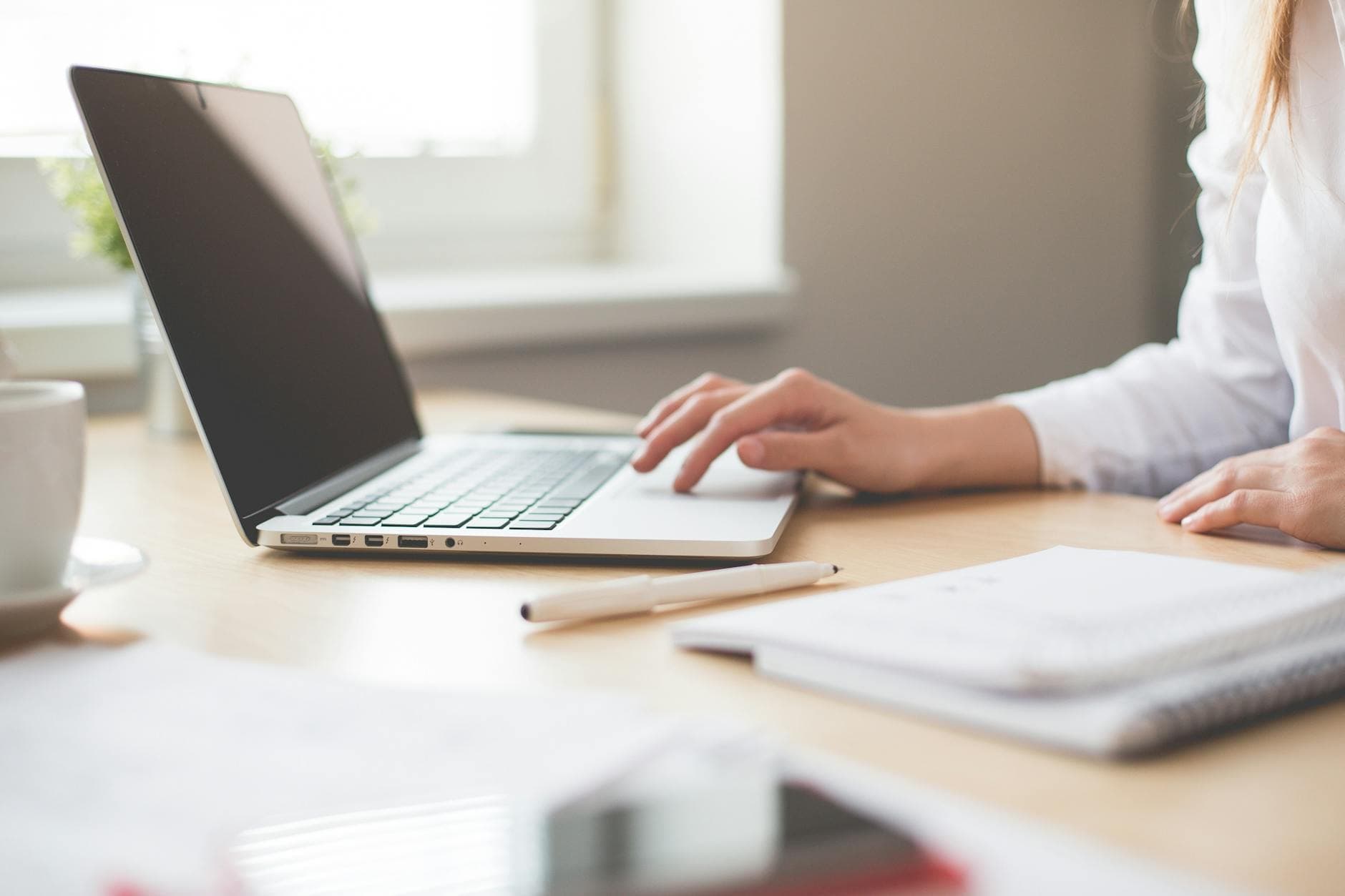 A person in a white shirt works on a laptop at a sunlit wooden desk, accompanied by a pen and a notebook. The clean, minimalist composition creates a professional and focused atmosphere.
