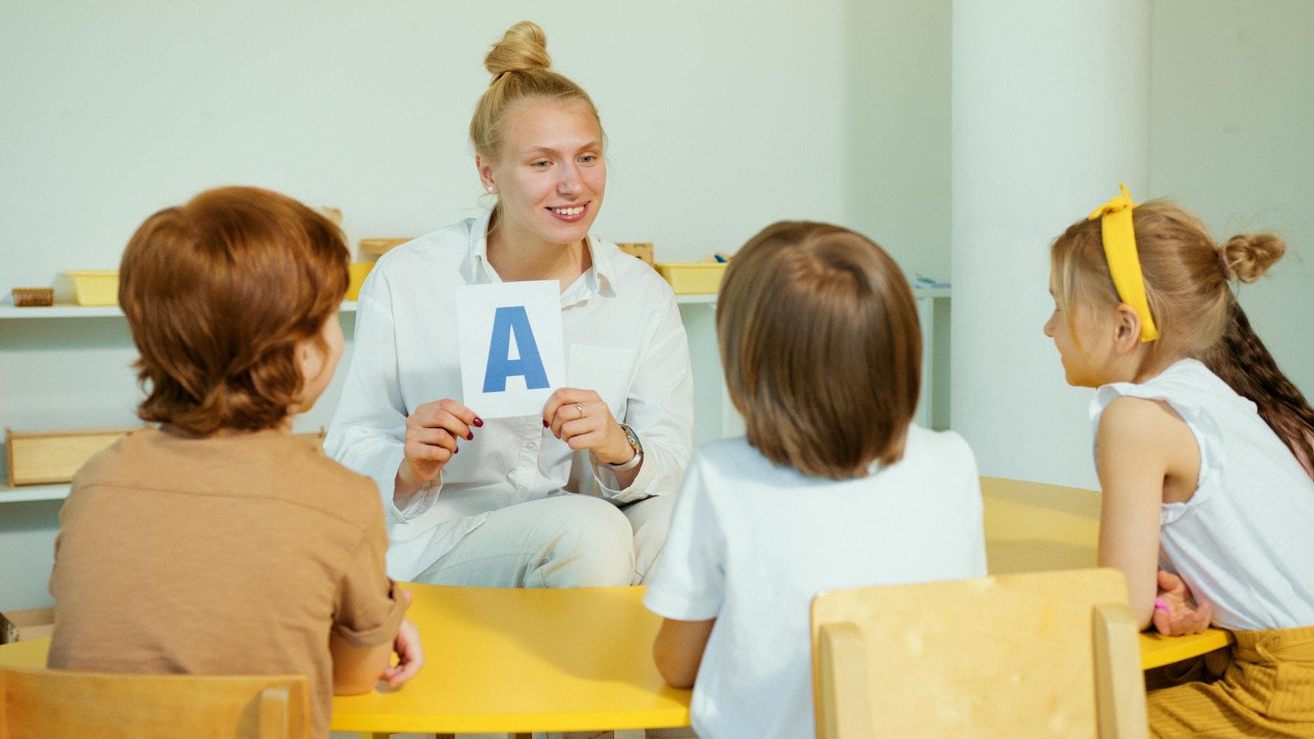 A teacher holds up a letter card while interacting with preschool children in a classroom setting.