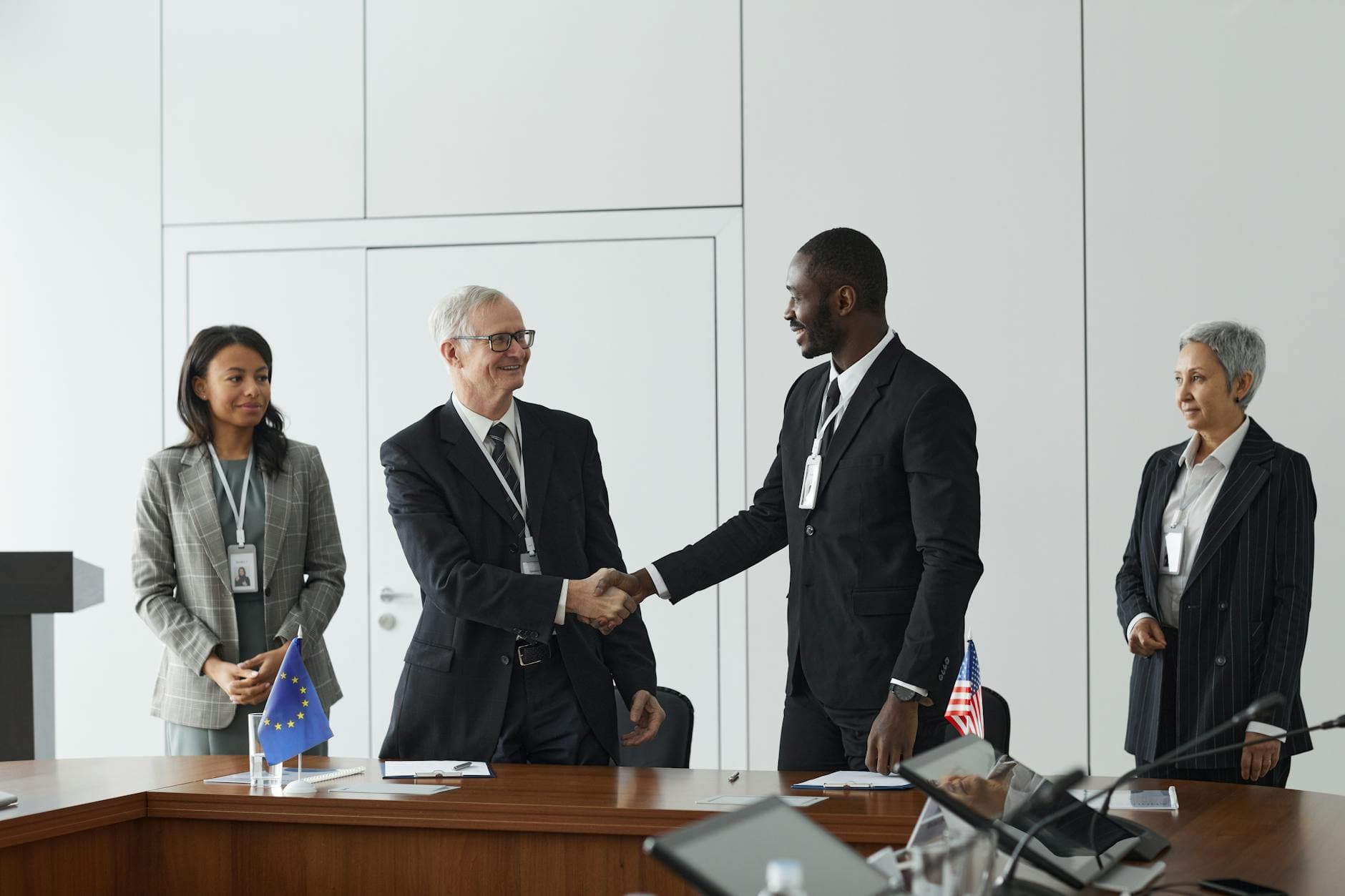 In a modern, minimalist conference room, two businessmen in suits shake hands, symbolizing an international agreement as indicated by the European Union and United States flags on the table. They are flanked by two female colleagues, all maintaining a professional and composed demeanor.
