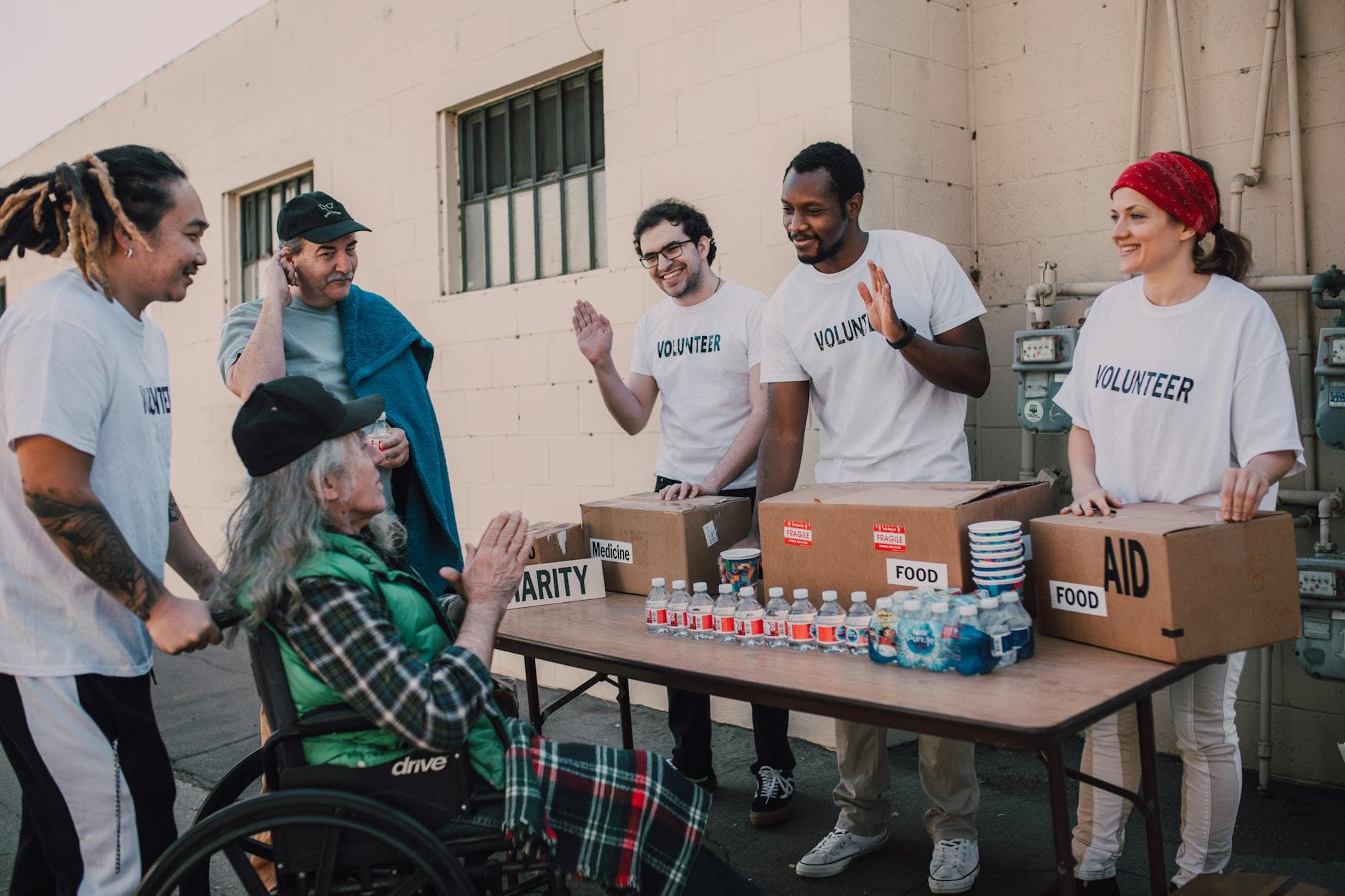 A group of diverse volunteers in matching white shirts interact warmly with an elderly person in a wheelchair and their companion at an outdoor aid station. The scene conveys a sense of community and support, with the volunteers offering food, medicine, and water from boxes arranged on a folding table against a simple wall.