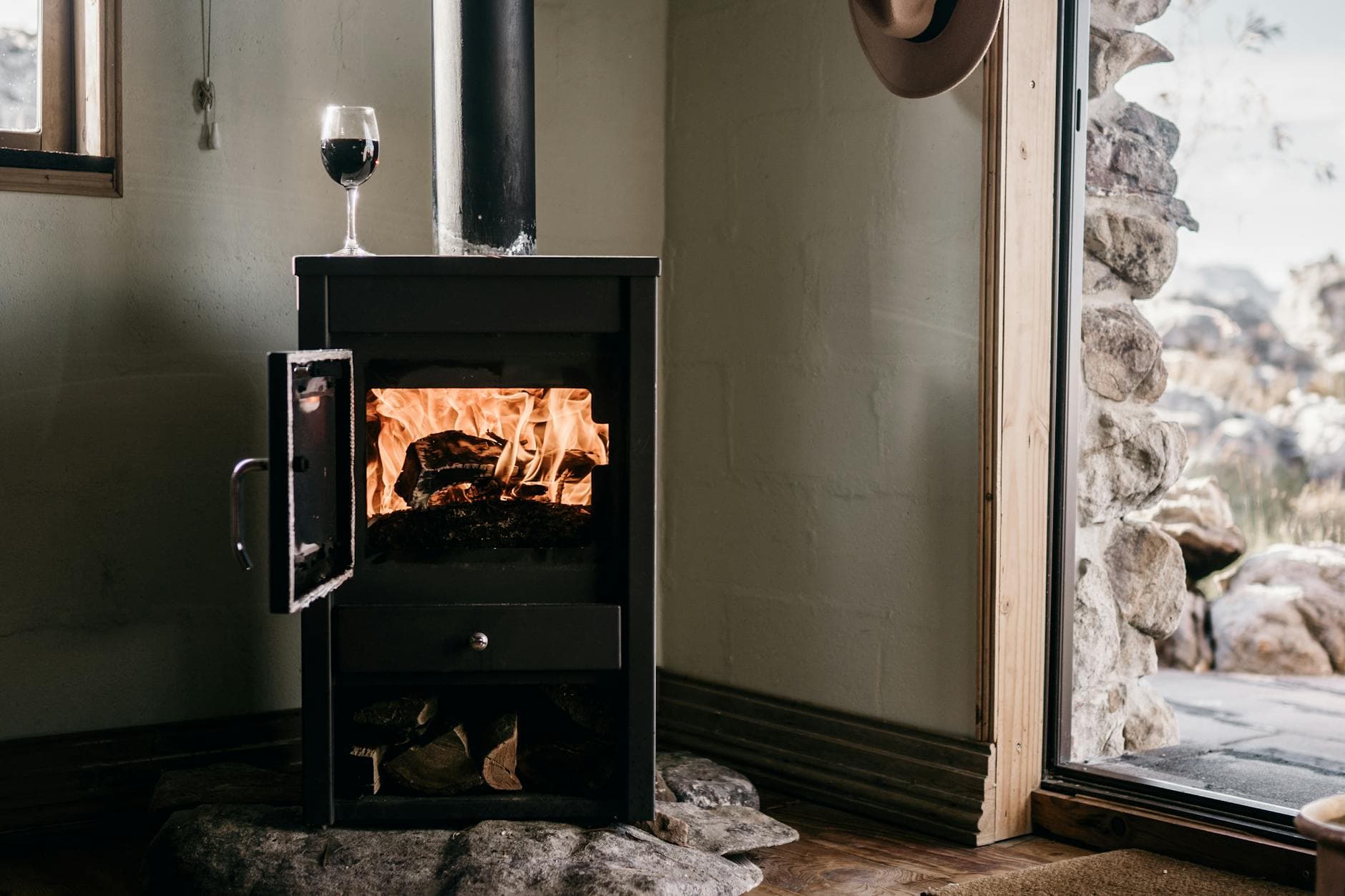 Warm indoor scene with a wood stove burning firewood beside an open door and glass of wine.