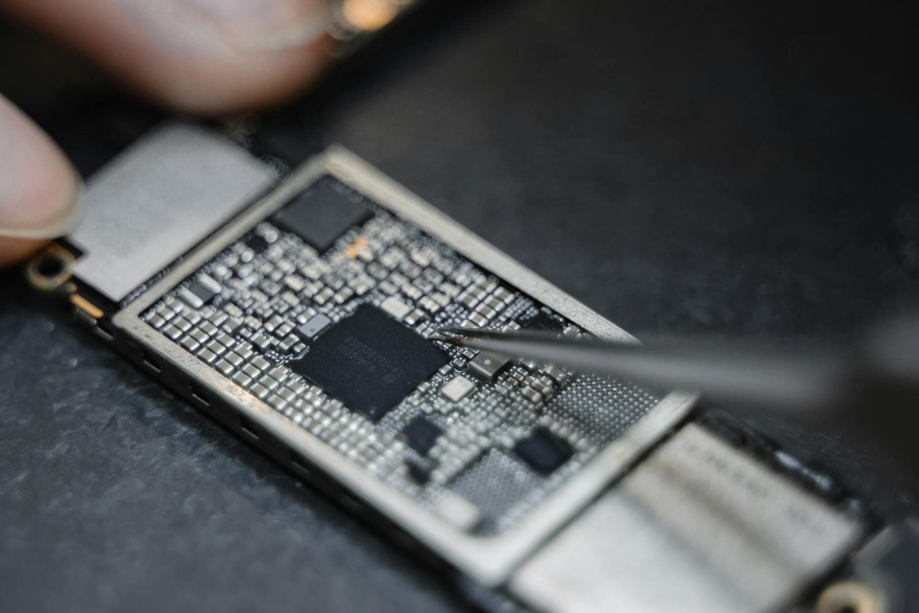 A technician’s hand uses metal tweezers to carefully manipulate a component on a complex electronic circuit board. The close-up shot emphasizes the intricate, industrial nature of device repair in a quiet, focused workspace.