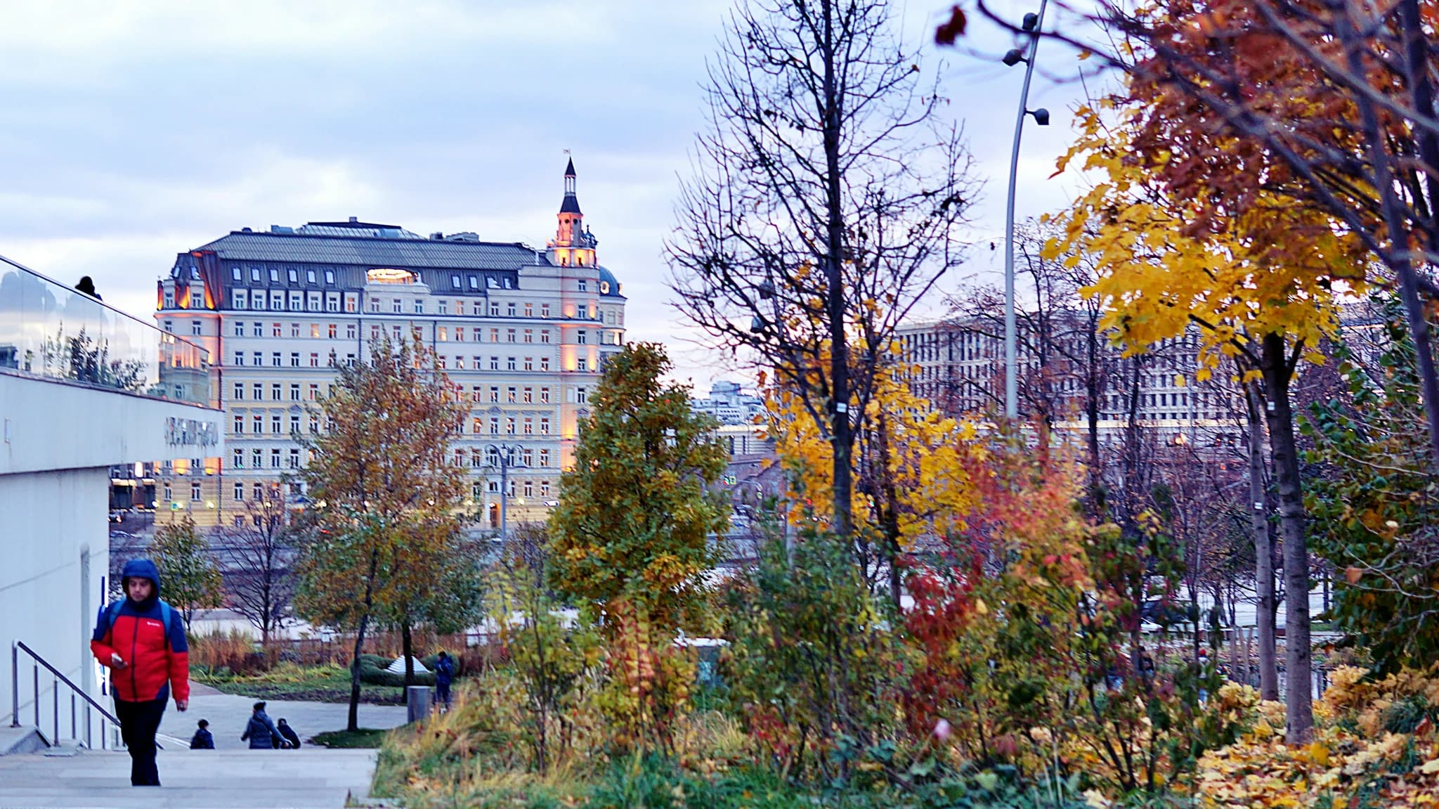 Person walking in park with autumn trees and building.