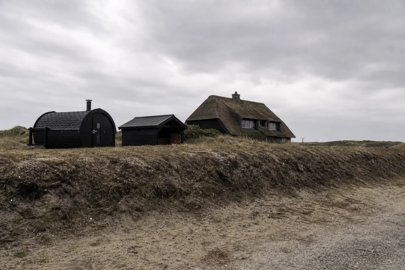 Træbeklædte småbygninger og jordvold i skellet ved et sommerhus.